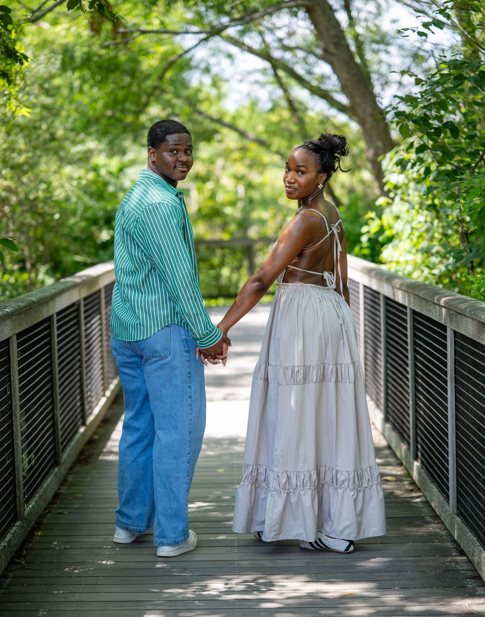 A couple holds hands on a wooden bridge surrounded by lush green trees, dressed in coordinating outfits.