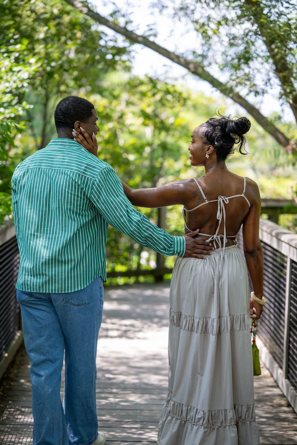 A Black couple walks hand-in-hand on a wooden bridge surrounded by lush green trees, dressed in elegant casual attire.