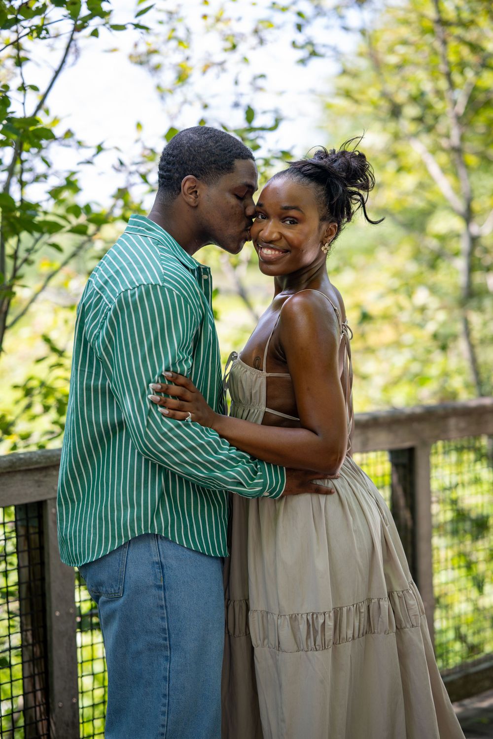 A young Black couple shares a romantic moment on a wooden bridge surrounded by lush green trees outdoors.