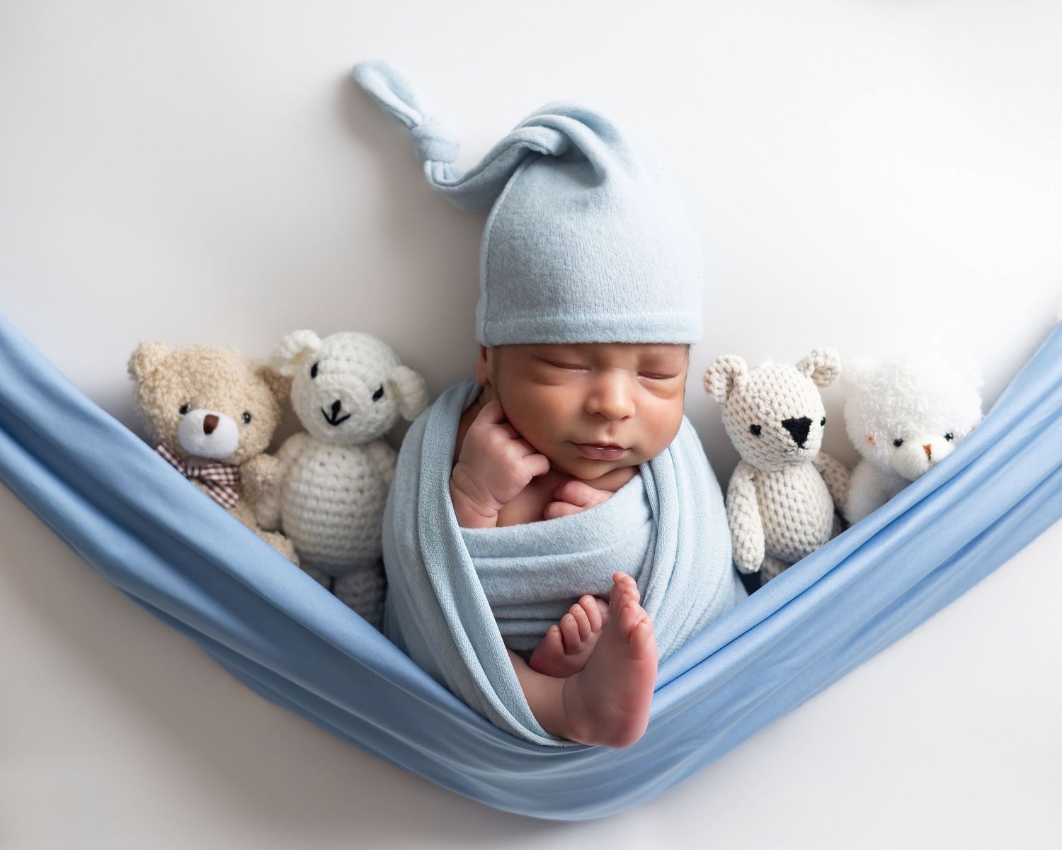Newborn baby wrapped in blue swaddle and knot hat, sleeping peacefully in fabric hammock surrounded by teddy bears.