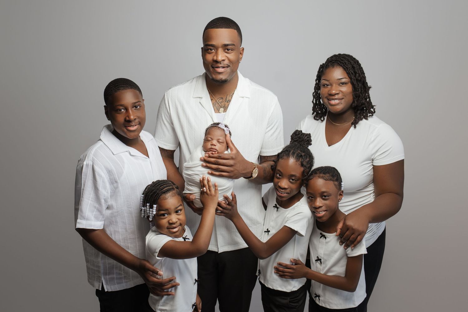 A joyful family portrait featuring parents and four children, with dad holding a newborn baby, all wearing white tops.