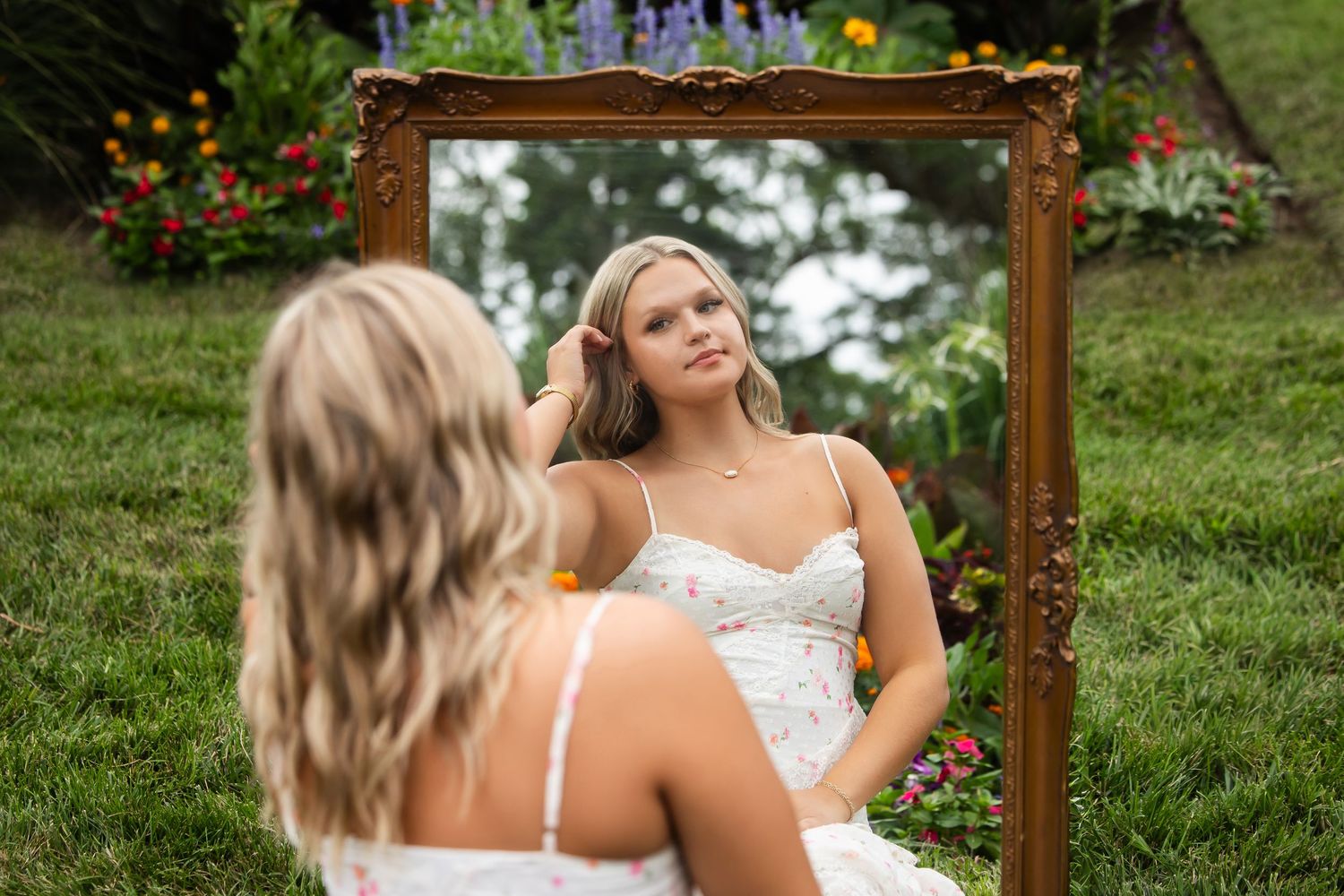 Young blonde woman in white floral dress sits outdoors examining her reflection in an ornate gold-framed mirror.