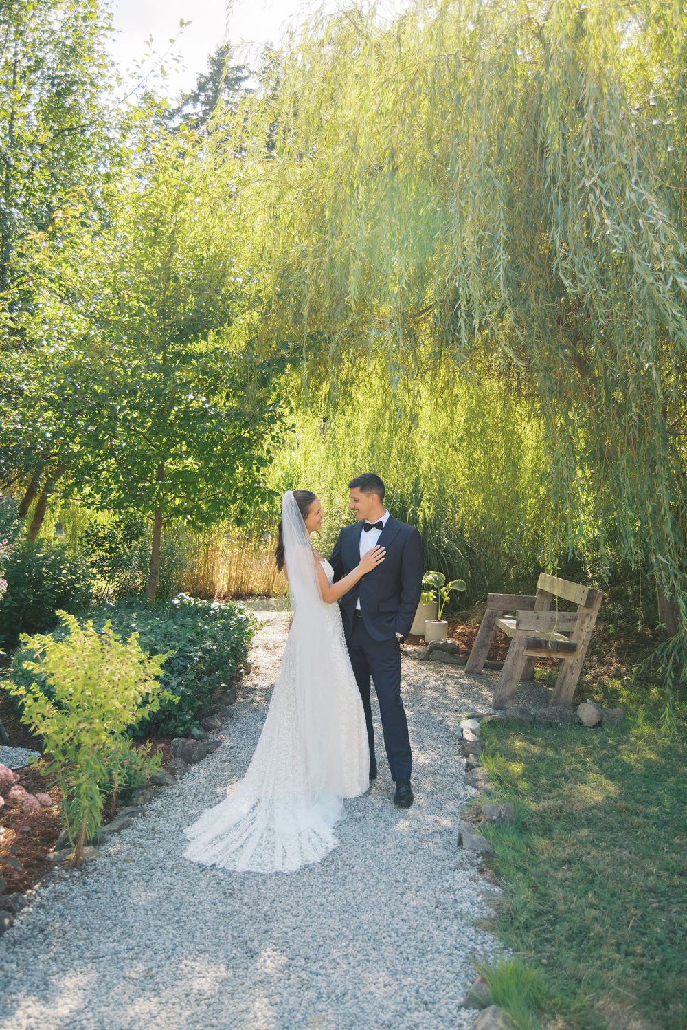 Bride and groom share a romantic moment on a gravel path beneath a weeping willow tree in a lush garden setting.