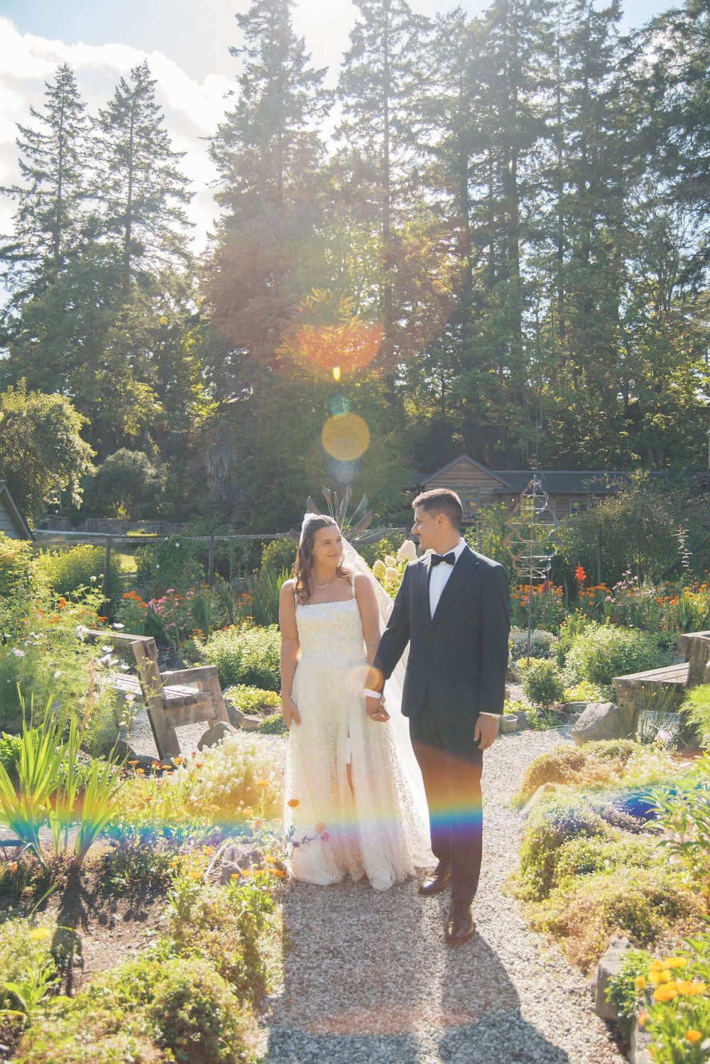 Bride and groom walk hand-in-hand through a sunlit garden path, surrounded by lush greenery and warm golden light.