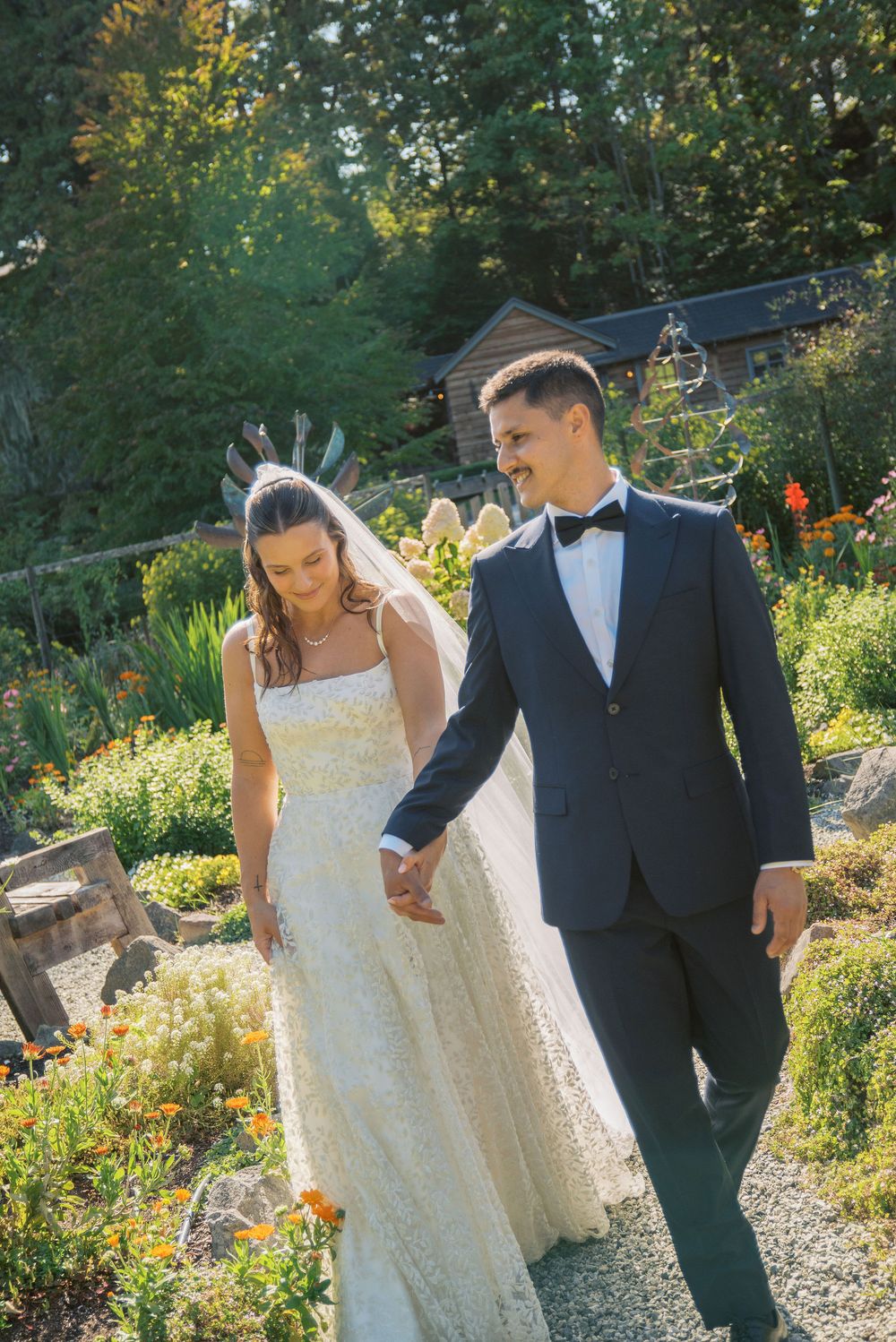 Bride and groom walk hand in hand through a sunlit garden, she in a lace gown, he in a navy tuxedo.