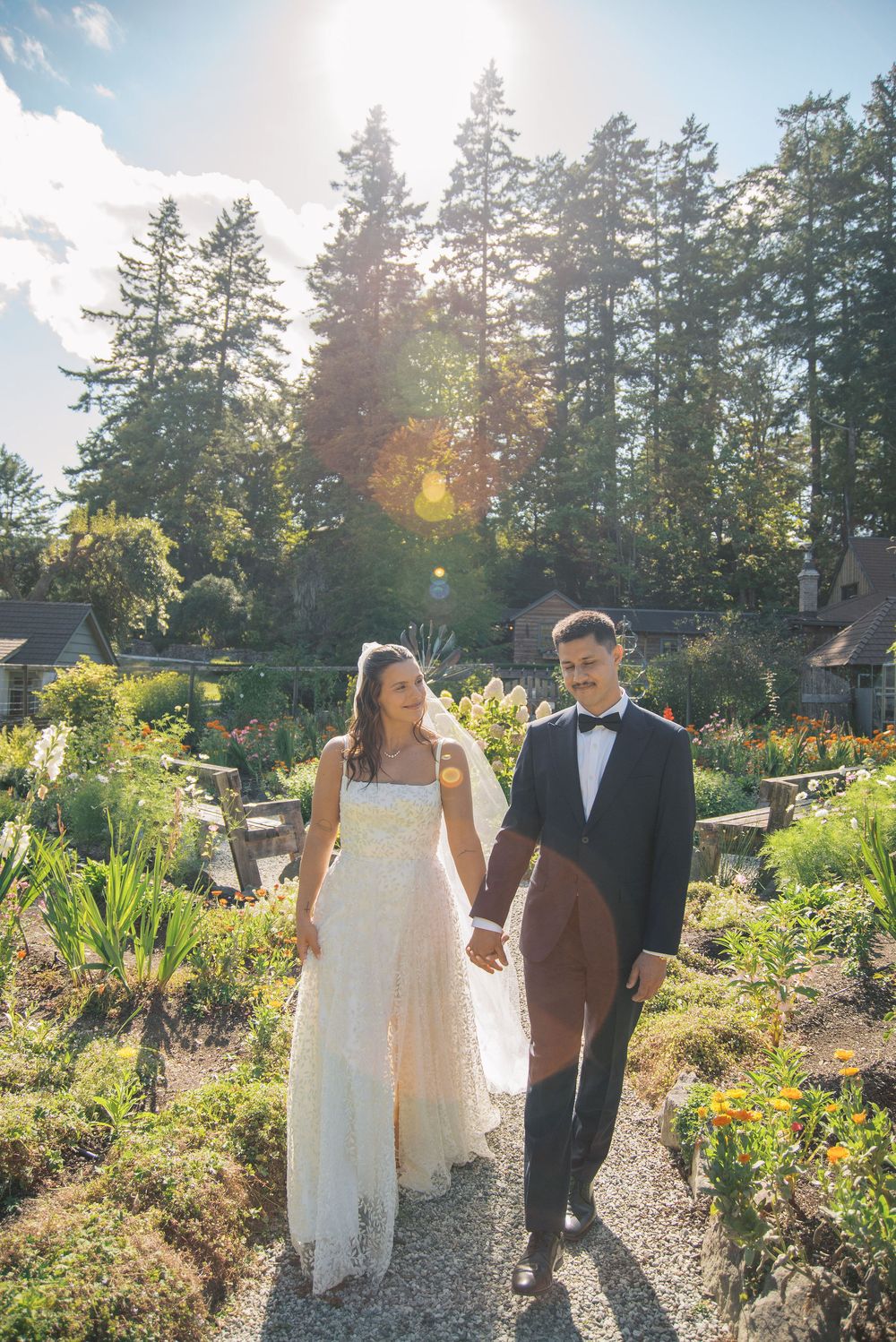 Bride and groom walk hand-in-hand through a sunlit garden path surrounded by lush greenery and tall trees.
