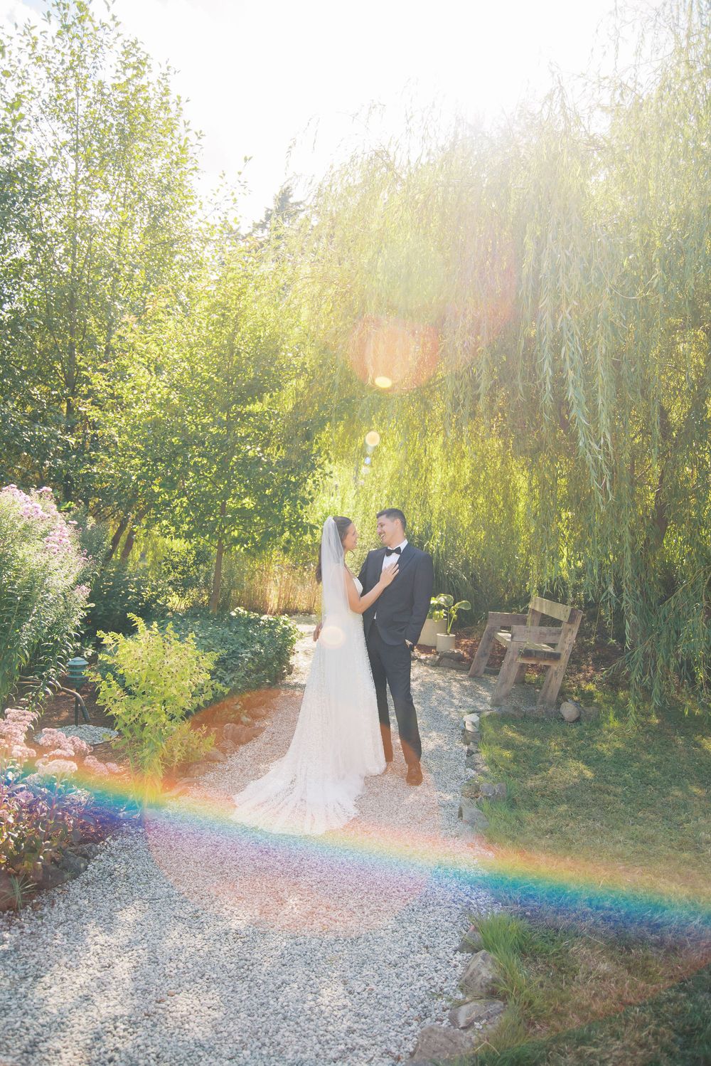Bride and groom share a romantic moment in a sunlit garden with a rainbow at their feet and a weeping willow backdrop.