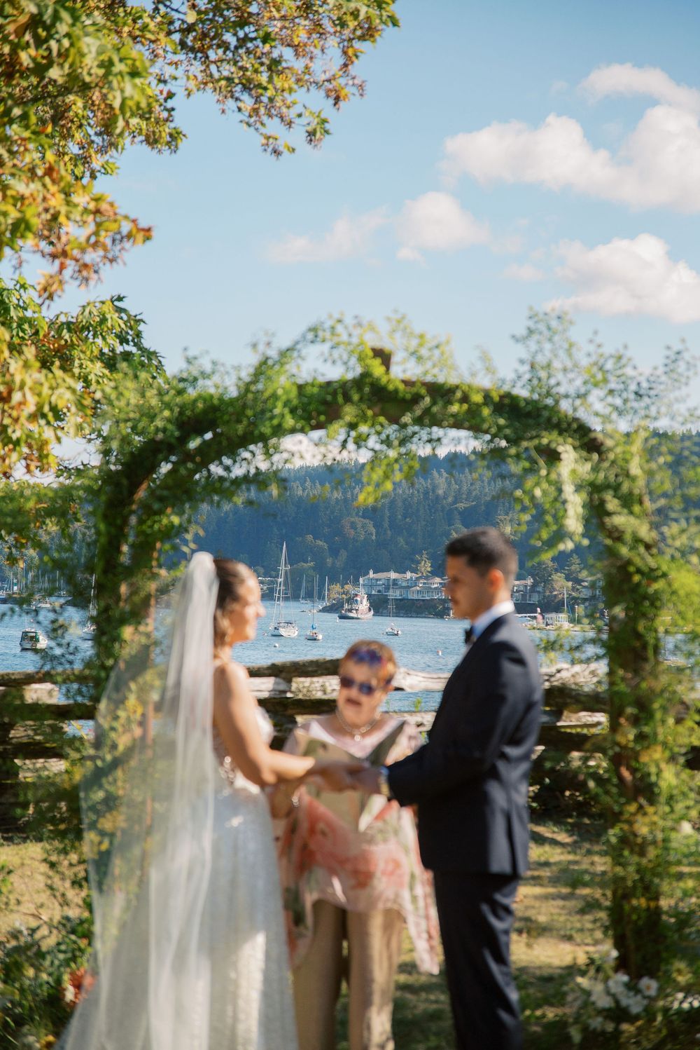 Couple exchanges vows under a floral arch with a scenic waterfront and sailboats in the background.