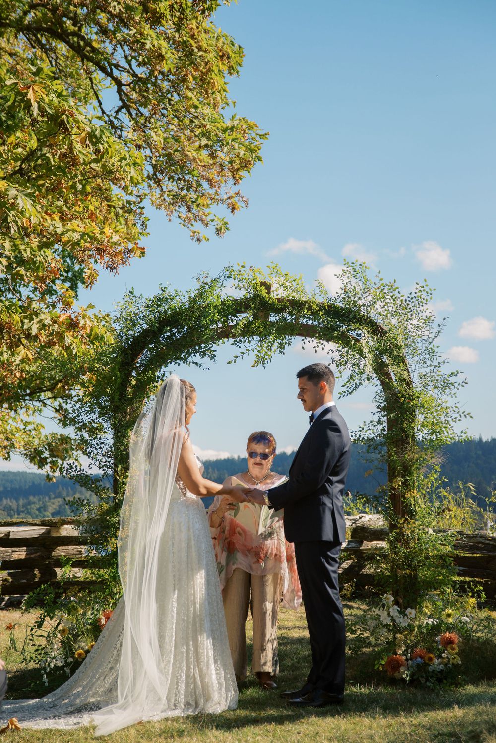 Bride and groom exchange vows under a lush green arch at an outdoor wedding ceremony on a sunny day.