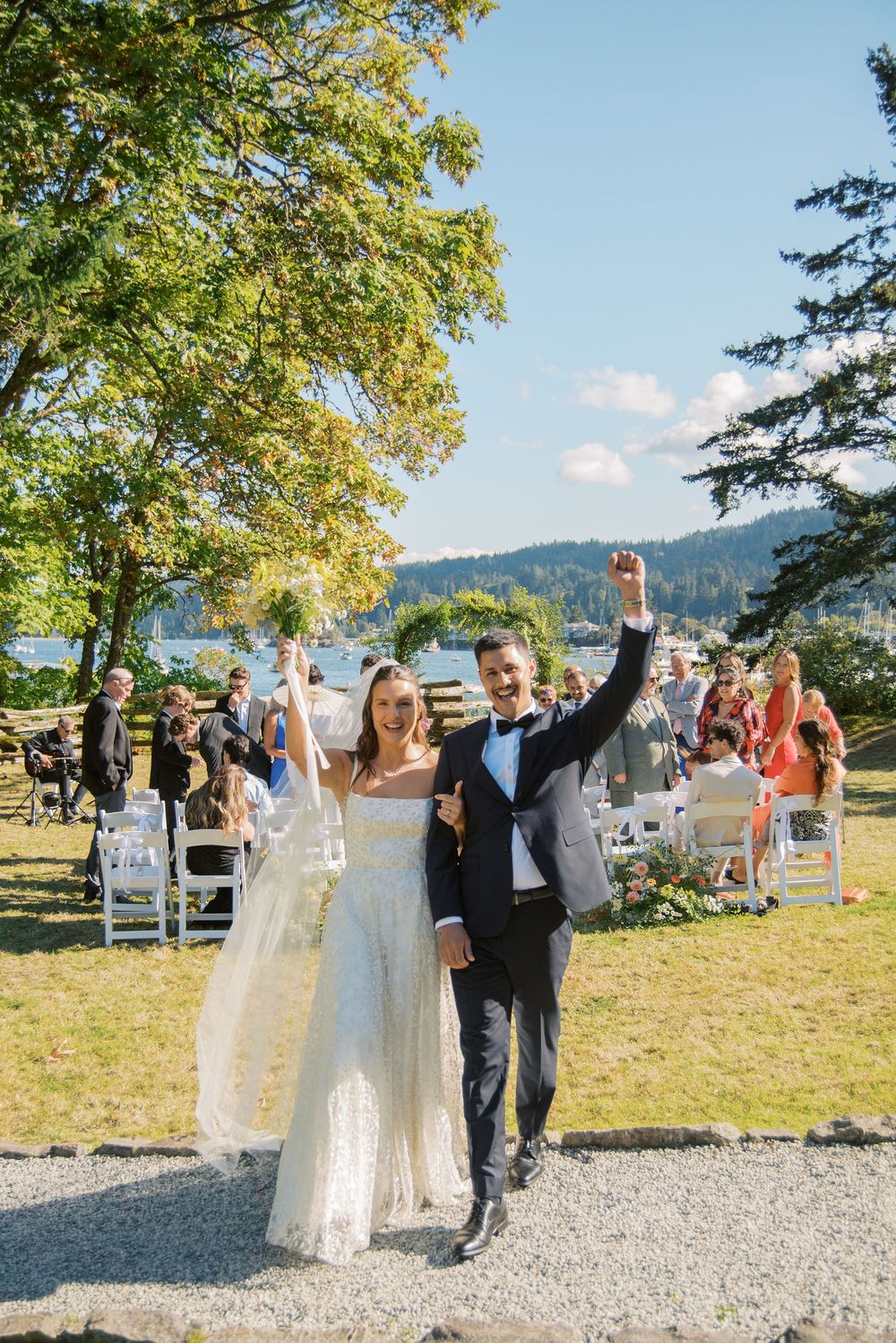 Joyful newlyweds raise their arms in celebration while walking down the aisle at an outdoor lakeside wedding ceremony.