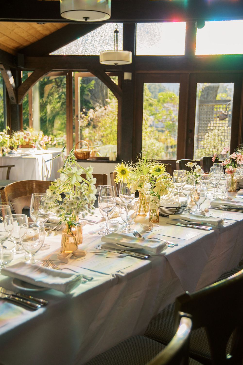 Sunlit wedding reception table with white linens, glassware, and sunflower centerpieces in a rustic venue.