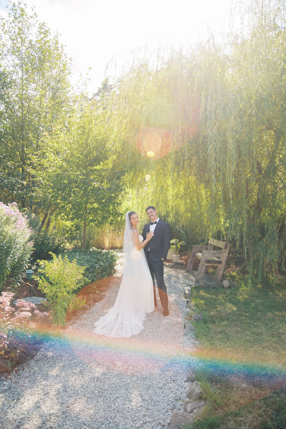 Bride and groom pose on a sunlit garden path with willow trees, lens flare, and a rainbow effect at their outdoor wedding.