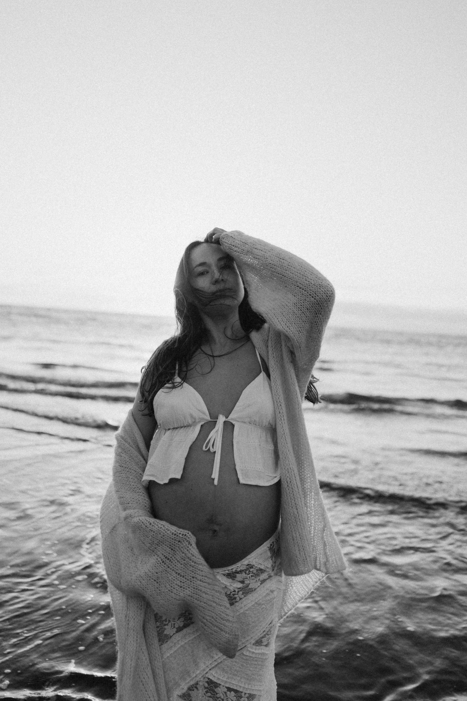 Pregnant woman in bikini top and knit cardigan poses gracefully on beach at dusk in black and white photo maternity session, Waikato.