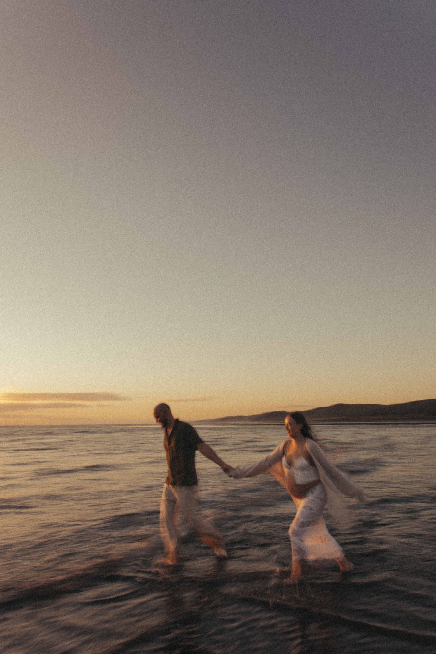 Couple holding hands walking in ocean waves at sunset with mountains in the background during a maternity session in New Zealand, Raglan.
