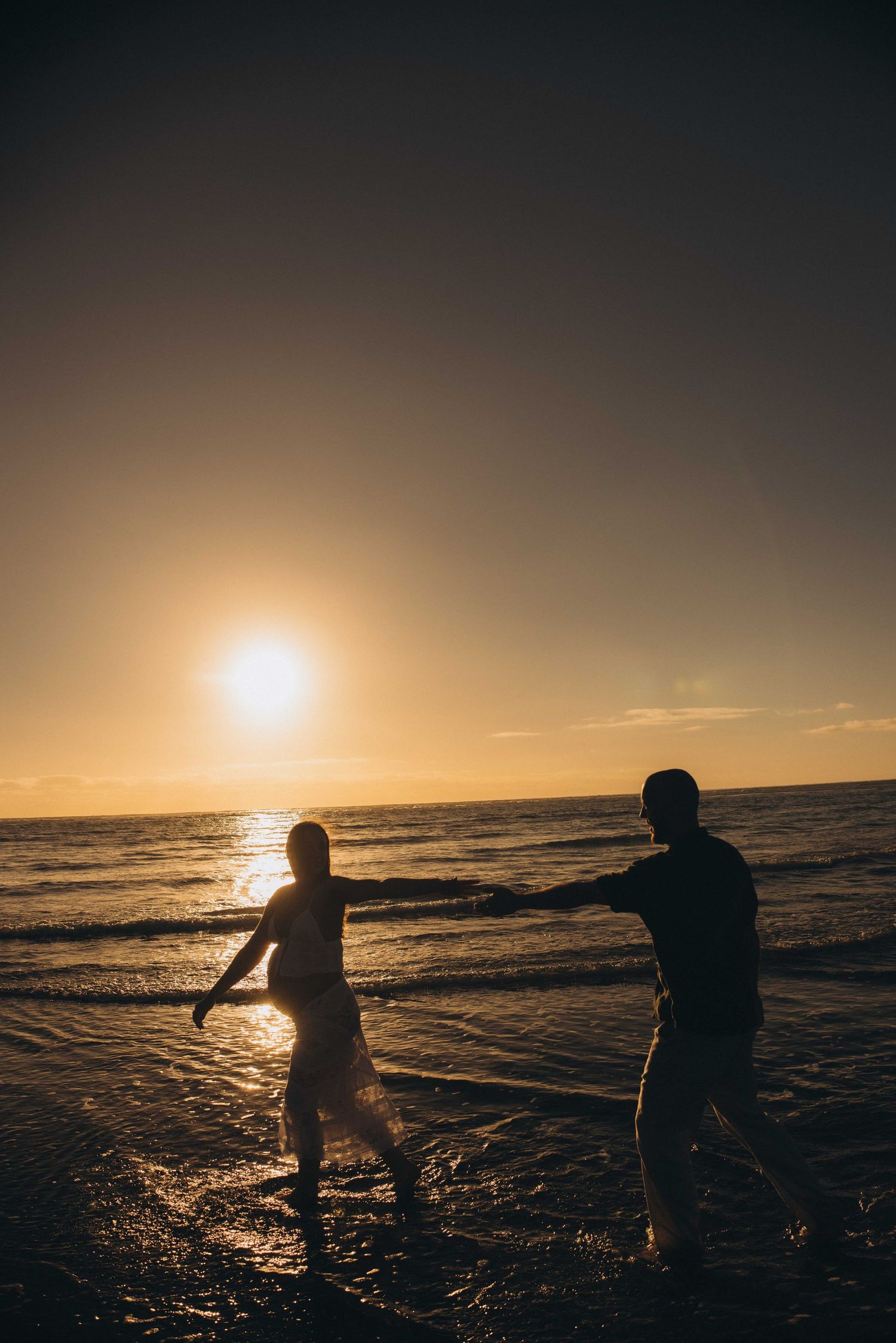 Silhouette of a couple holding hands and walking on a beach at golden sunset with ocean during a maternity session in New Zealand, Raglan.