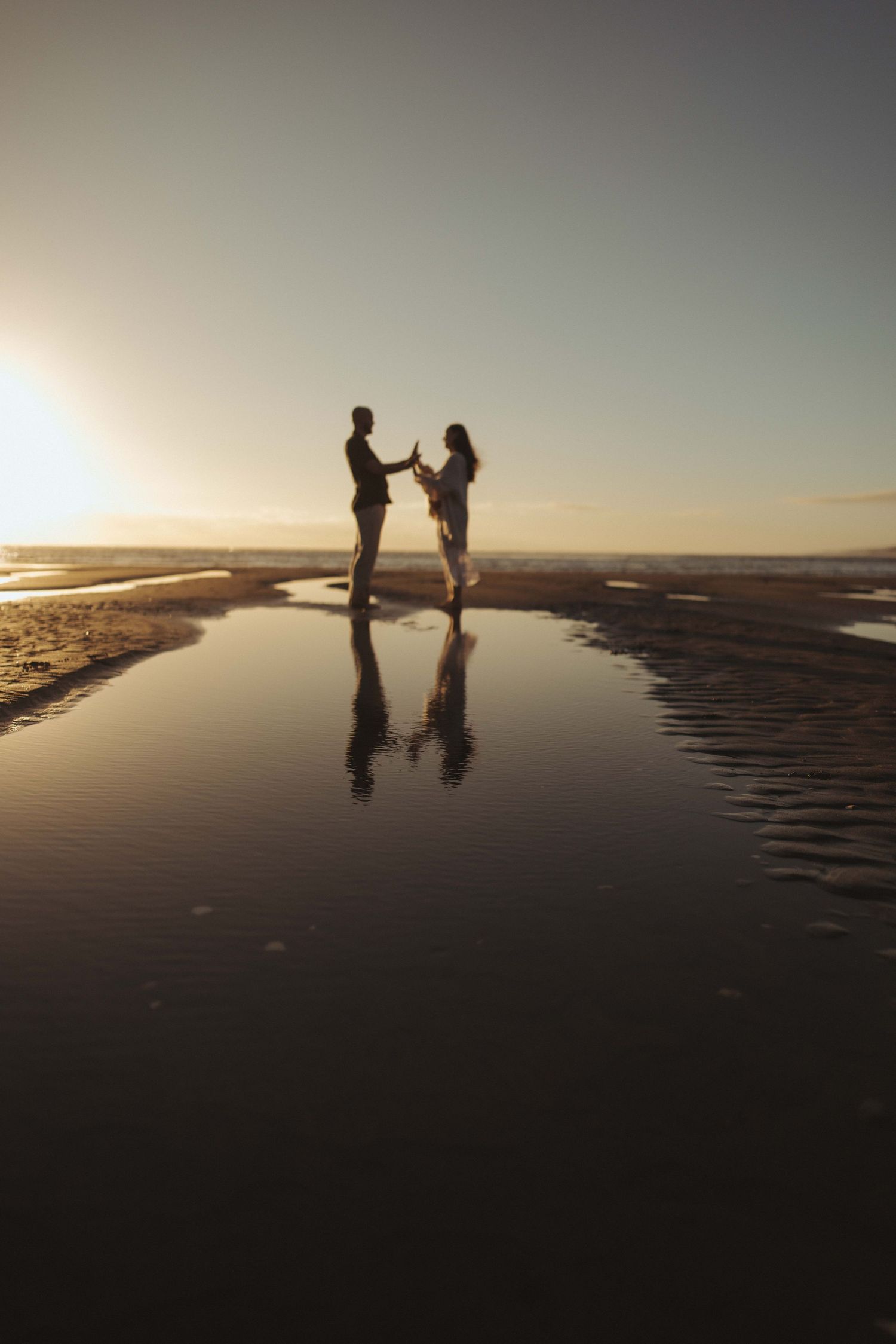 Couple holding hands at sunset on a reflective beach, silhouetted against golden sky with mirror-like water reflection.