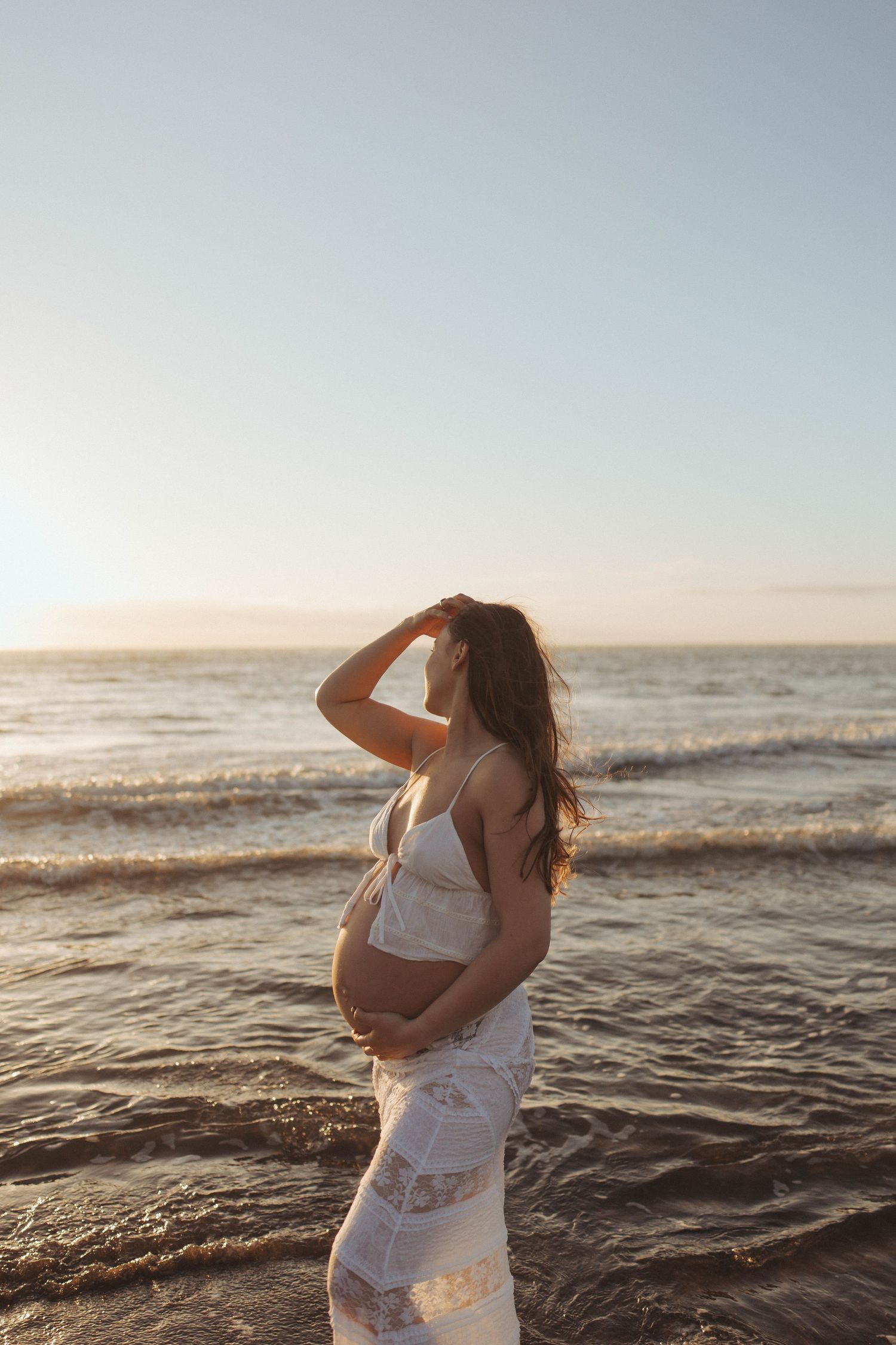 Pregnant woman in white lace dress cradling her belly at sunset on a serene beach with ocean waves in Raglan New Zealand.