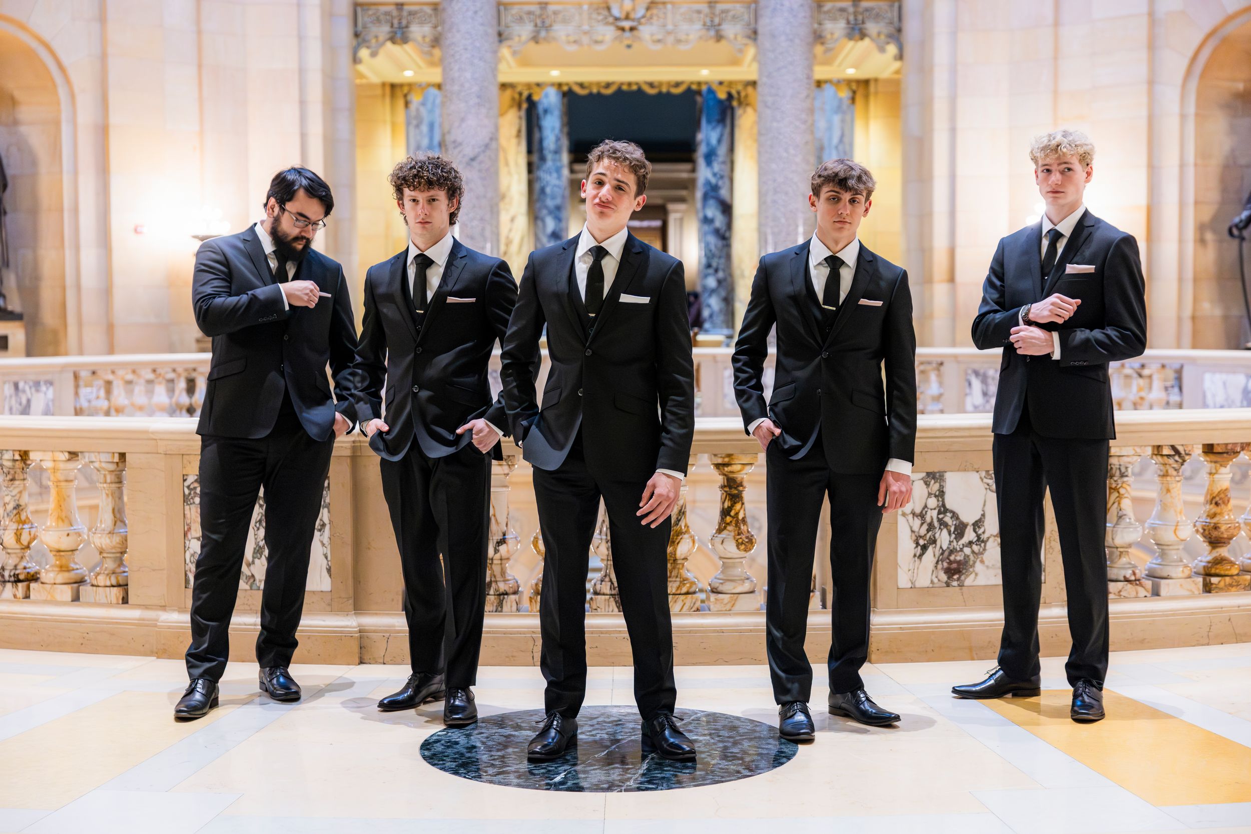 Five young men in matching black suits pose confidently in an elegant marble interior.