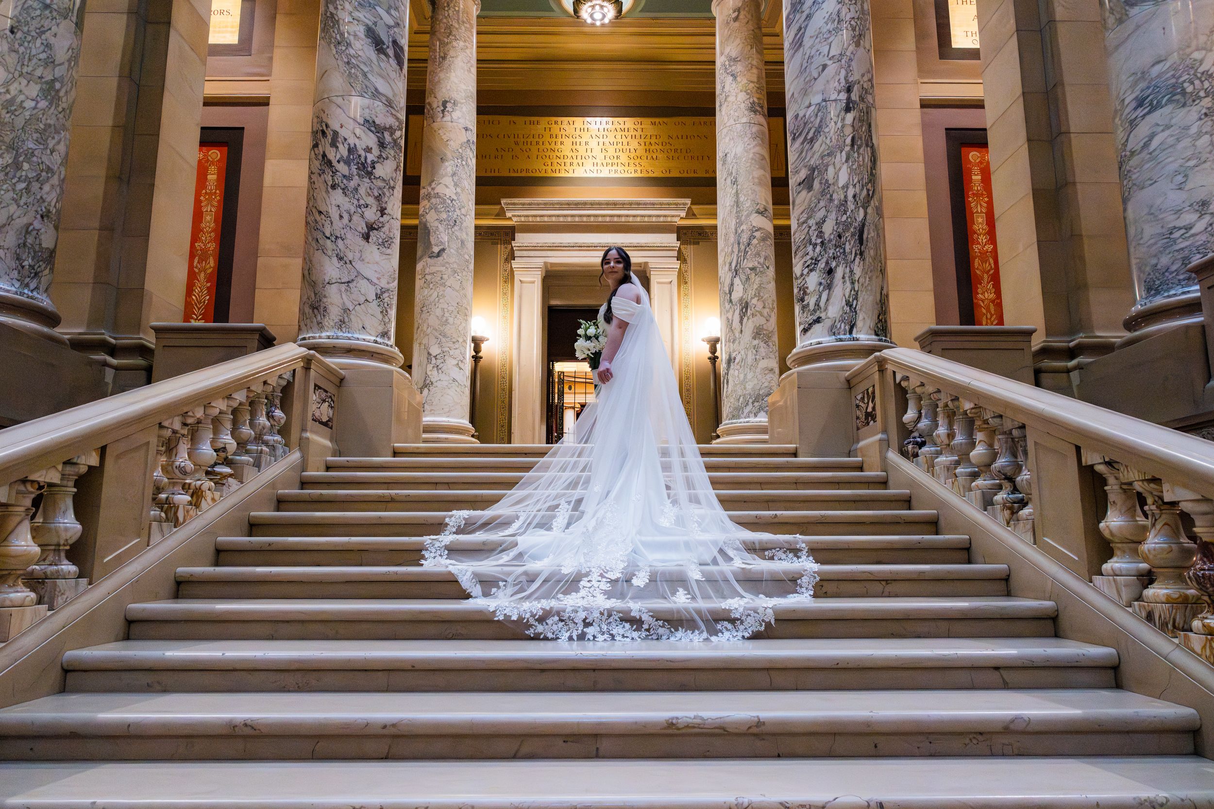 Bride in stunning white gown with long lace cathedral veil poses on grand marble staircase.