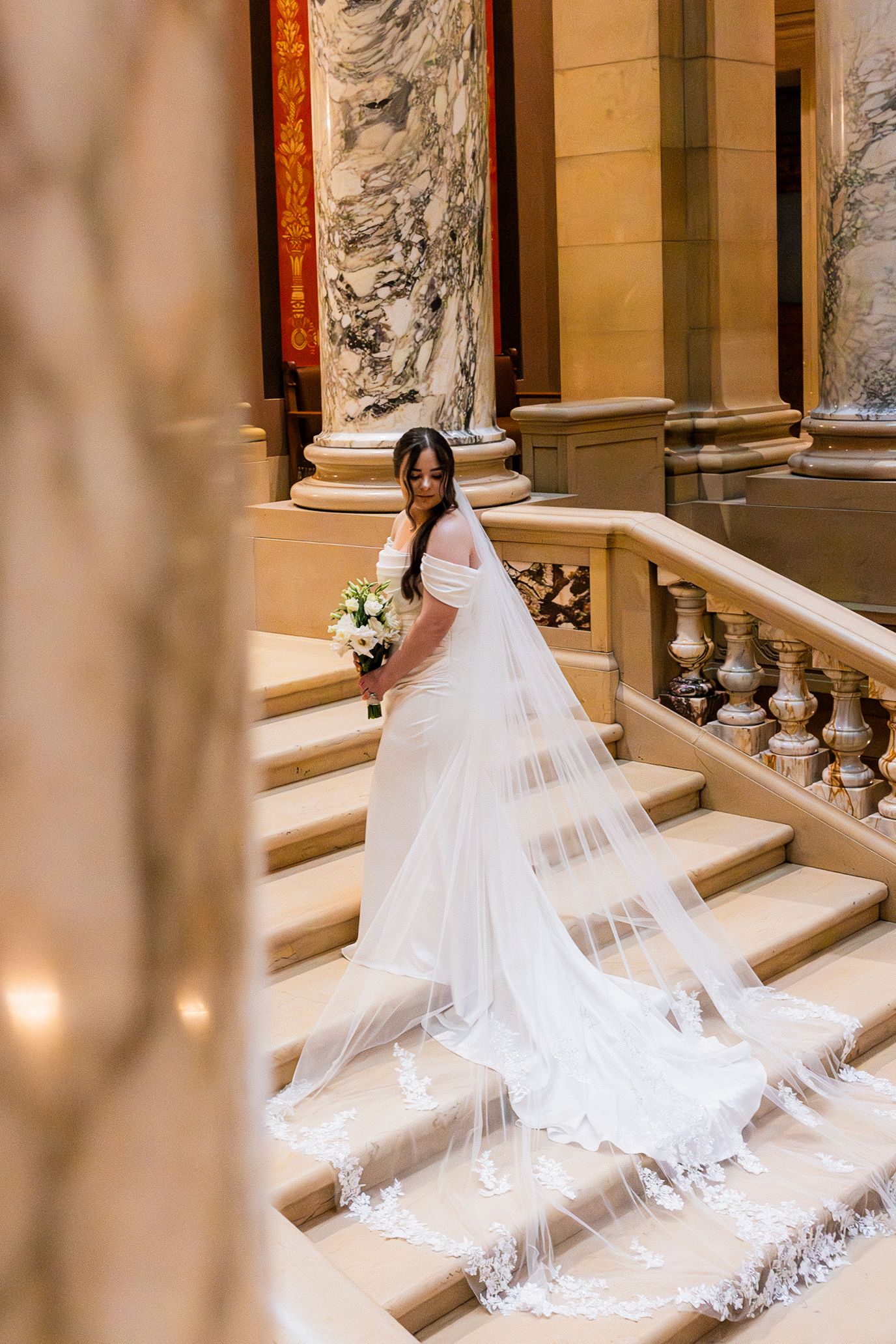 Bride in white gown with long lace veil descends marble staircase holding bouquet in elegant columned venue.