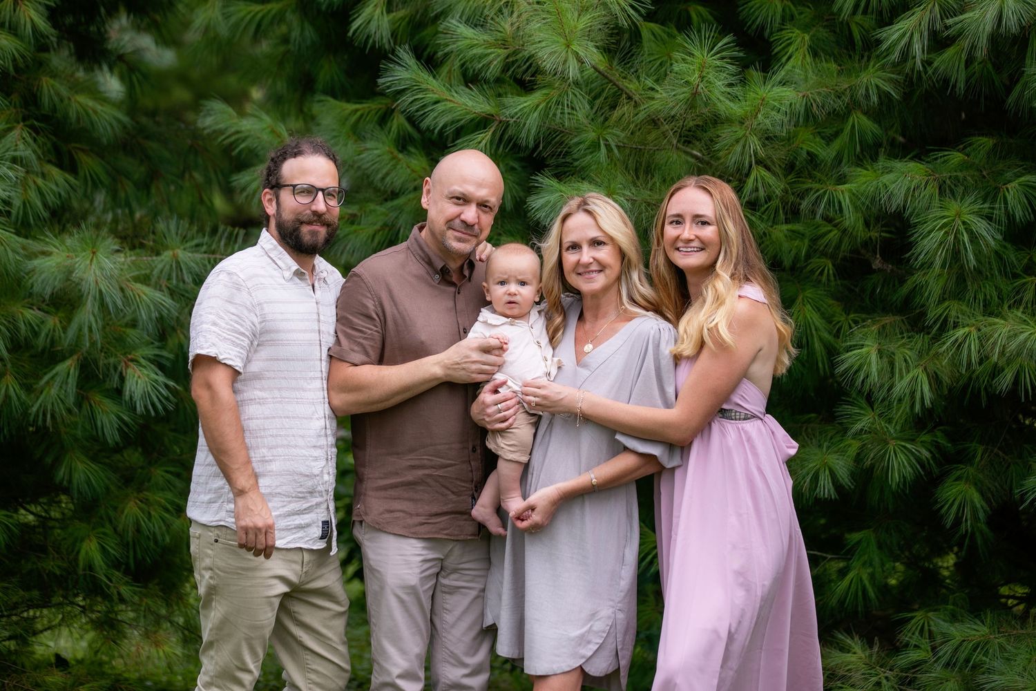 Family of five poses outdoors in front of lush green pine trees, including a baby held by parents.