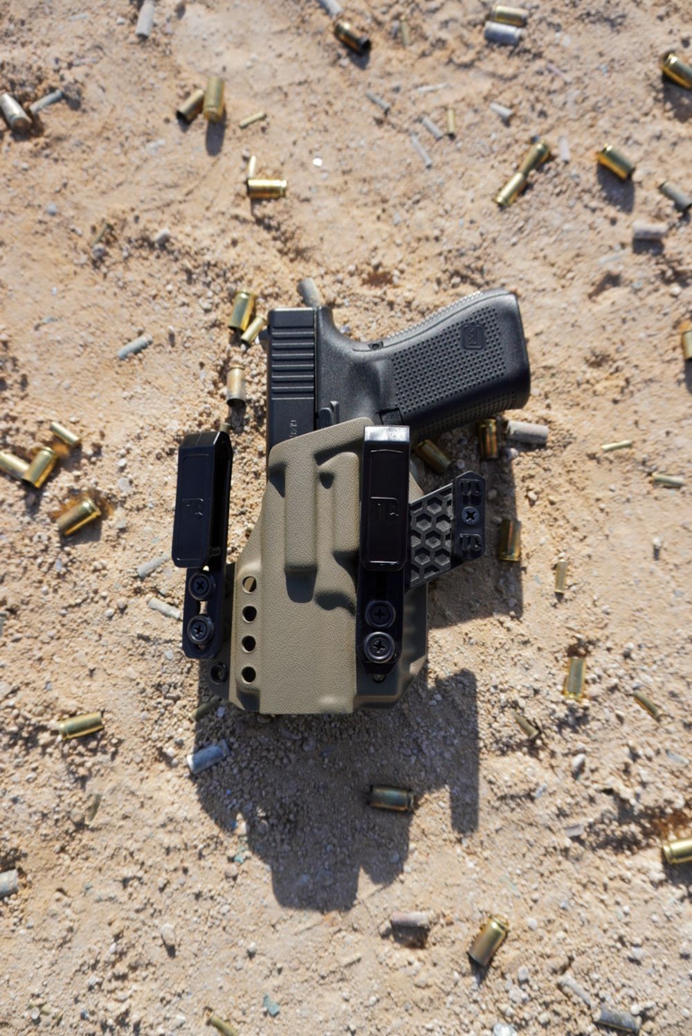 A Glock pistol in an olive drab holster surrounded by spent brass casings on sandy ground at a shooting range.