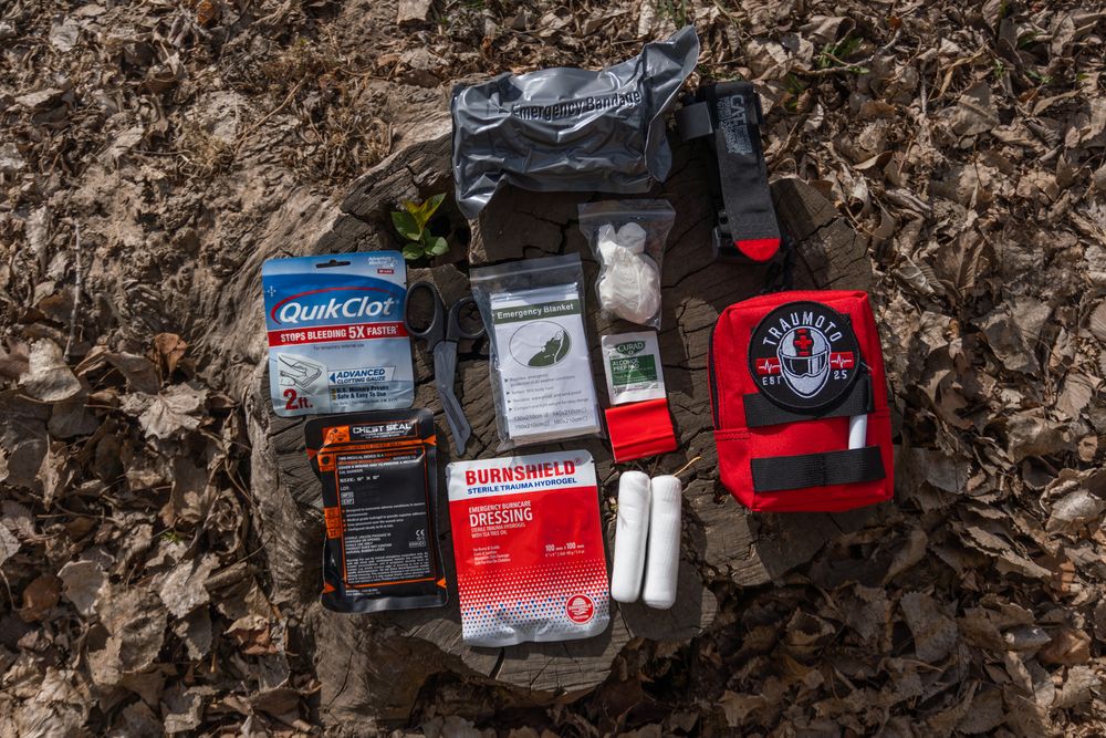 First aid kit contents including QuikClot, tourniquet, bandages, and emergency supplies laid out on fallen leaves.
