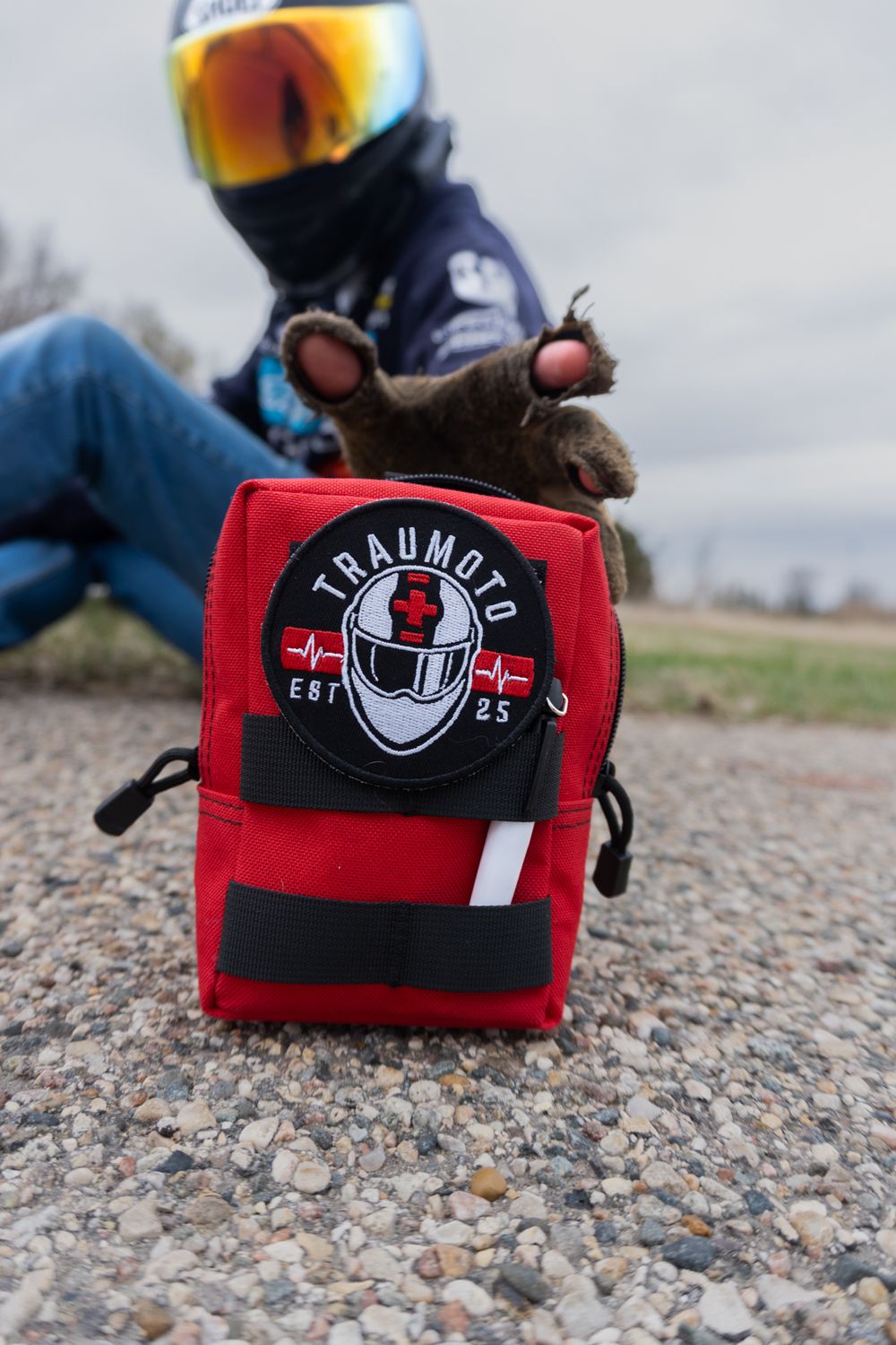 Red Traumoto motorcycle first aid kit bag on gravel with helmeted rider in background.