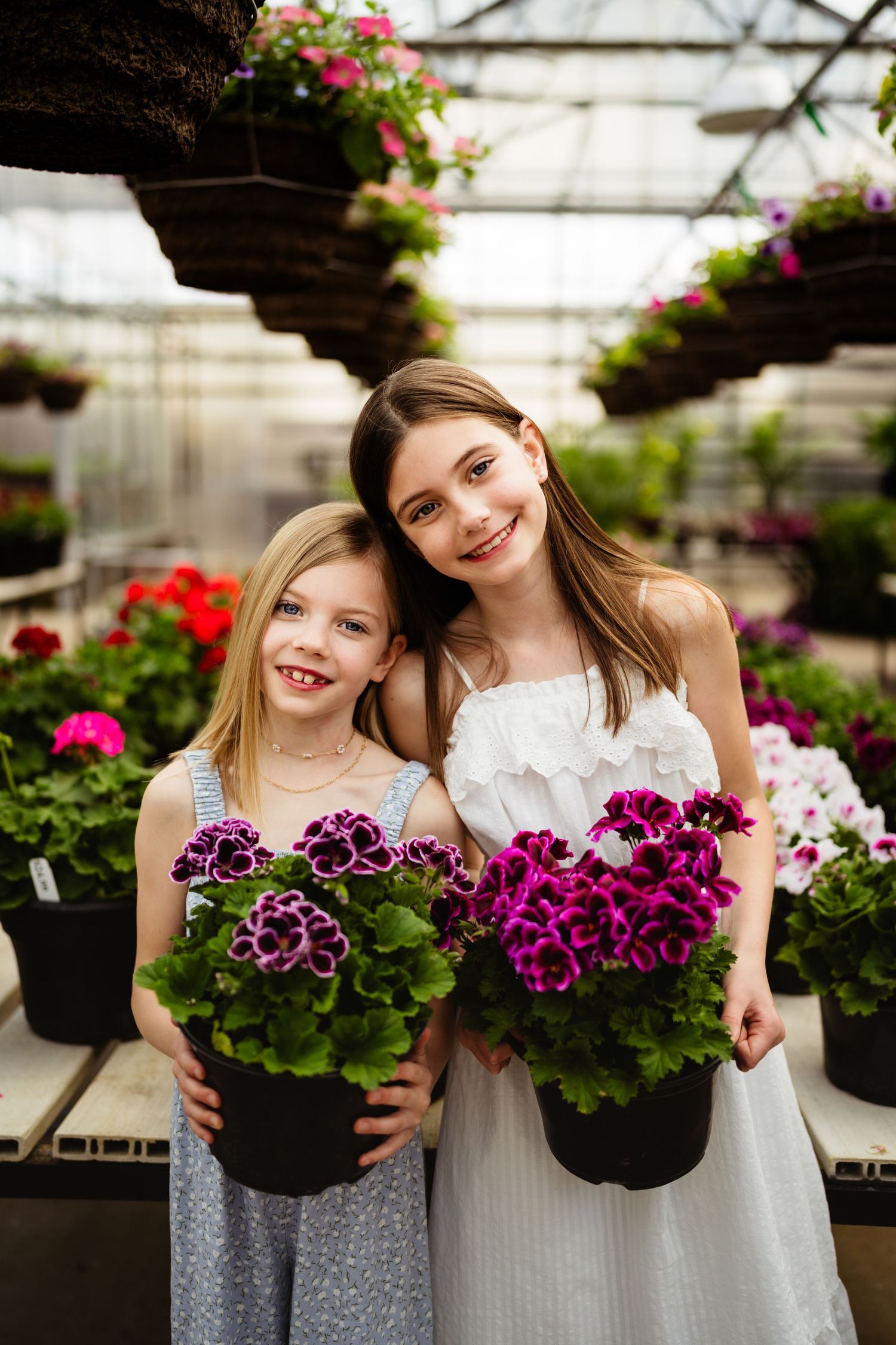 Two smiling girls holding purple flowering potted geraniums in a bright greenhouse garden center.