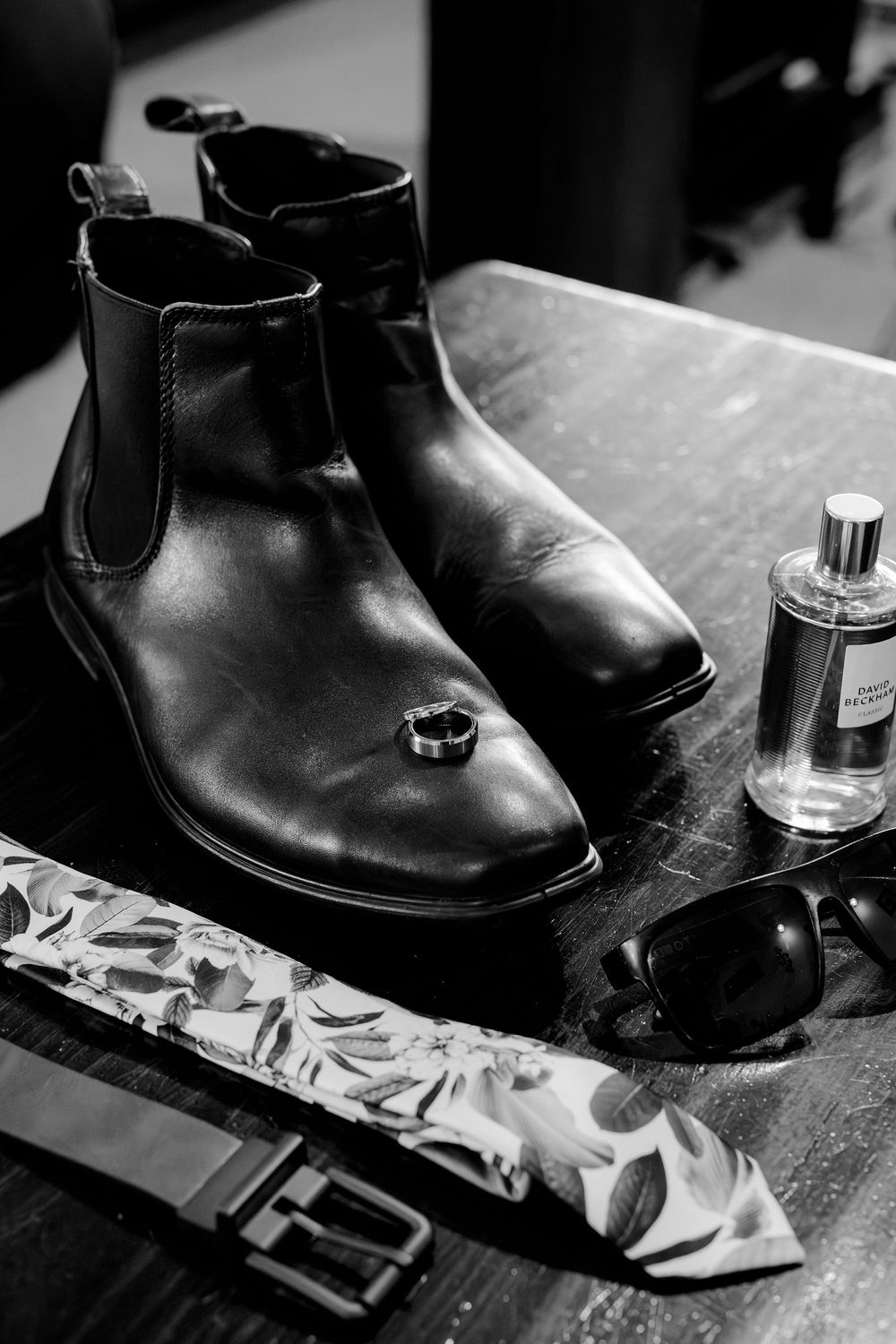 Black and white photo of mens wedding accessories including Chelsea boots, a ring, cologne, sunglasses, tie, and belt on a wooden table.