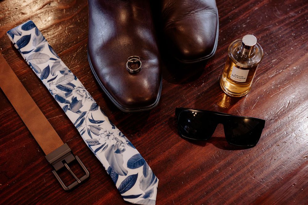 Groom accessories flat lay featuring brown leather shoes, floral tie, belt, sunglasses, cologne, and gold ring on wood floor.