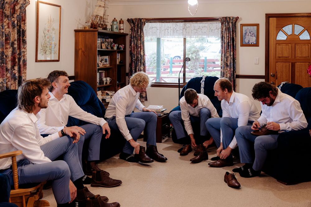 Groomsmen in white shirts and blue pants getting ready together in a cozy living room, putting on dress shoes before a wedding.