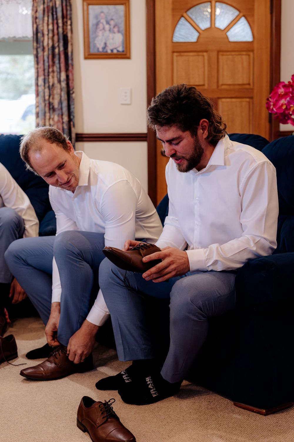 Two groomsmen in white shirts and blue trousers putting on brown dress shoes before a wedding ceremony.