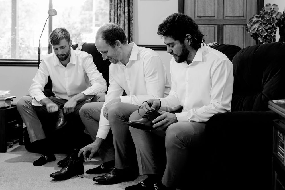 Three men in white shirts sit together getting dressed, adjusting shoes and cuffs in a candid black and white photo.