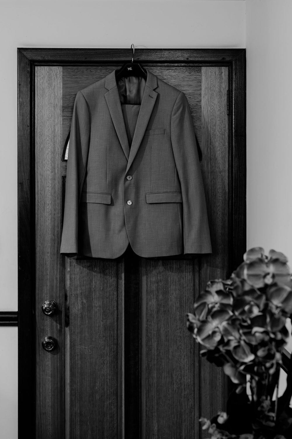 Black and white photo of a suit jacket hanging on a dark wooden door beside a floral arrangement.