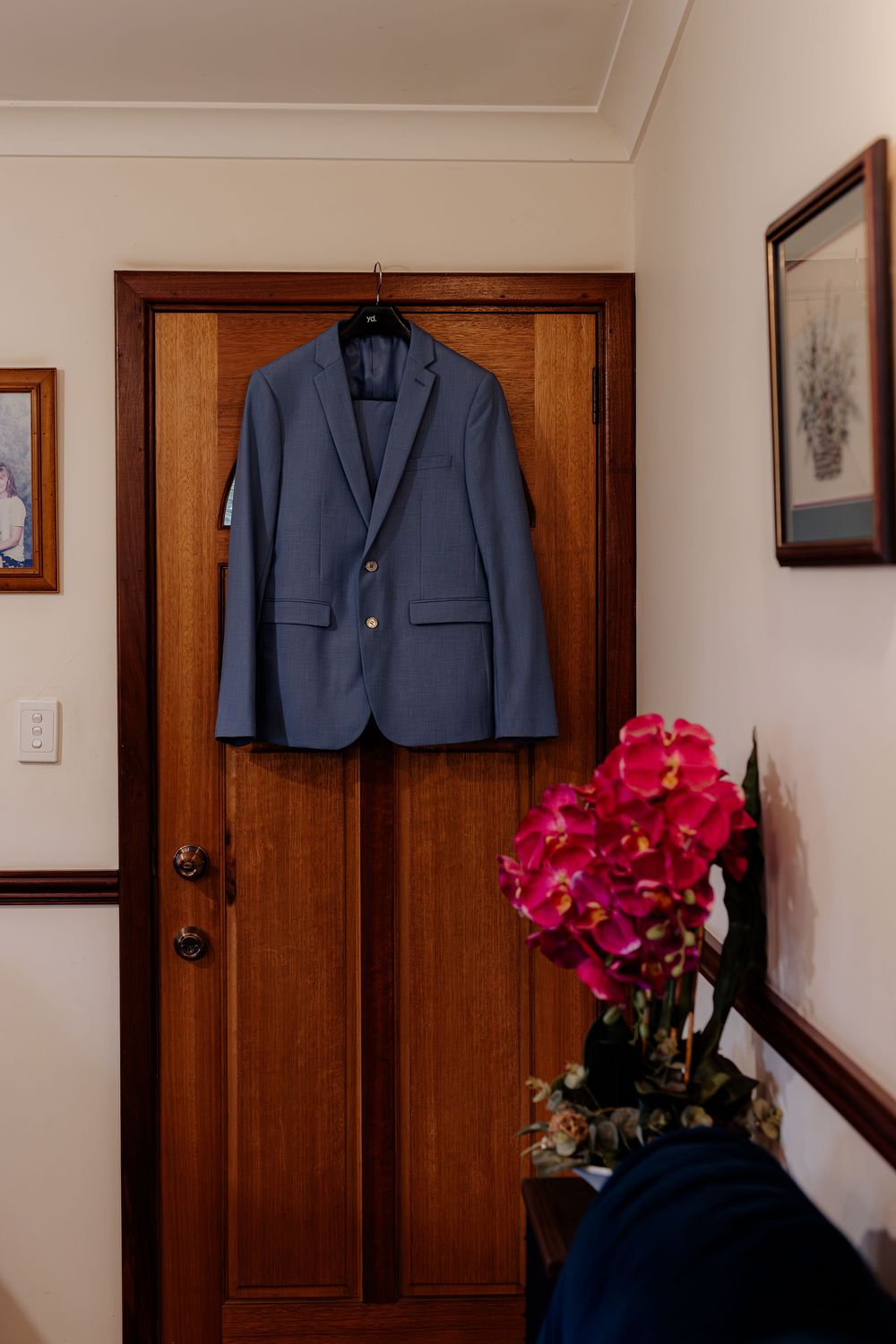 Blue suit jacket hanging on wooden door hanger beside pink flowers in a home hallway.
