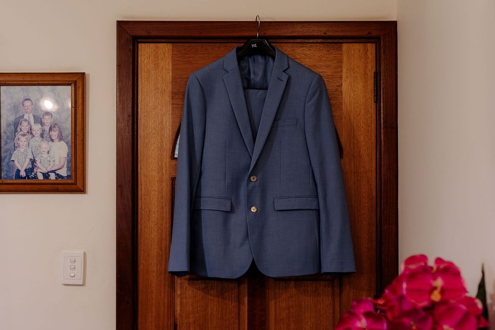 Navy blue men's suit jacket hanging on a wooden door with a family photo and pink flowers nearby.