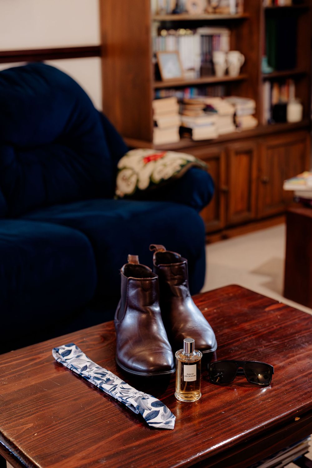 Brown leather Chelsea boots, floral tie, cologne, and sunglasses arranged on a wooden table in a cozy study.