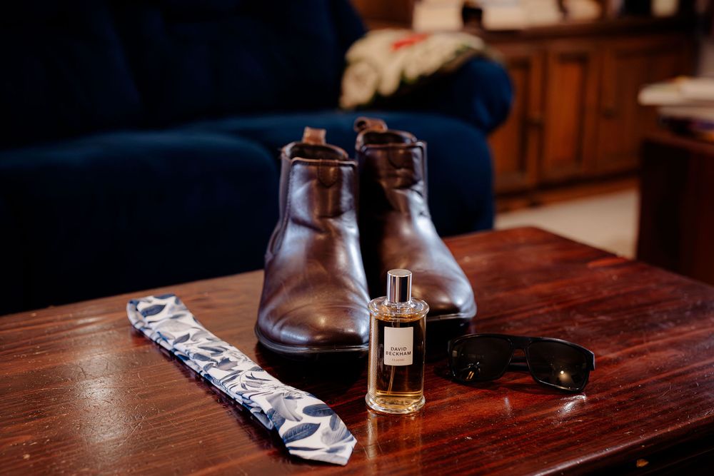 Men's brown leather dress boots, patterned tie, cologne bottle, and sunglasses on a wooden table.