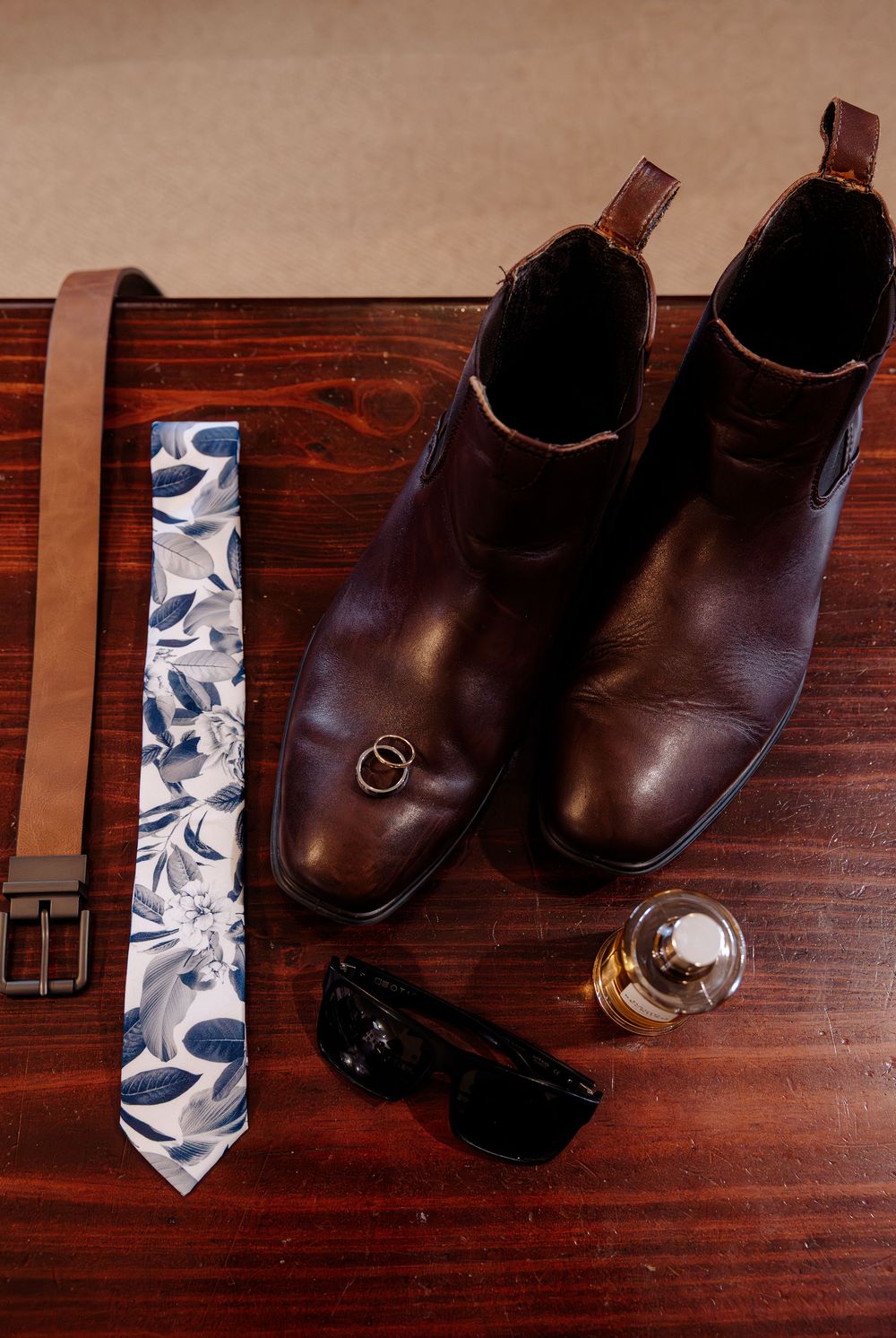 Brown leather Chelsea boots with floral tie, belt, sunglasses, gold rings, and cologne on wooden surface.