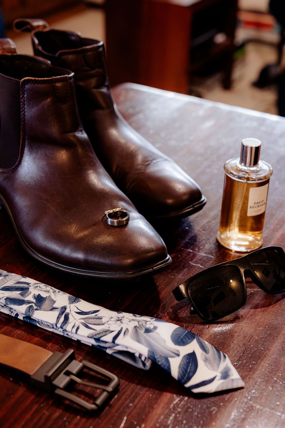 Dark brown Chelsea boots, cologne, sunglasses, floral tie, and belt arranged on a wooden table for a groom.