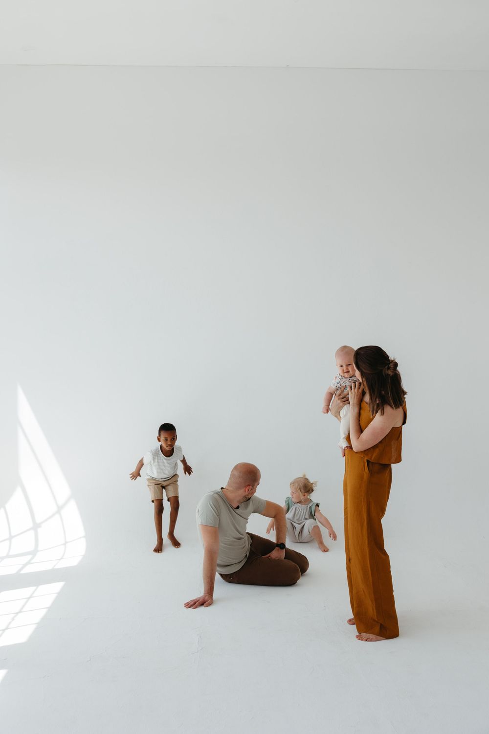 Family with three children posing in bright white studio, mother holding baby while father sits on floor.