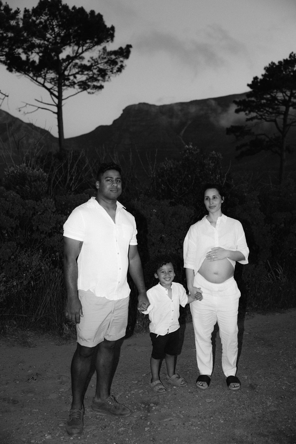 Black and white family maternity photo with parents and child in white outfits, mountains in background.