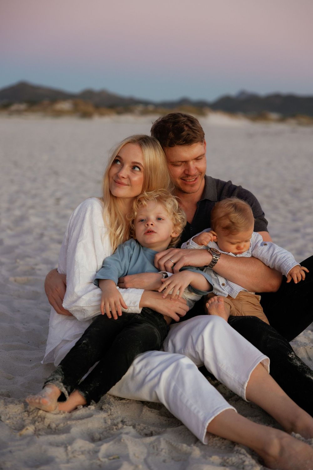 Family of four sitting on sandy beach at sunset, parents smiling with two young children in warm golden light.