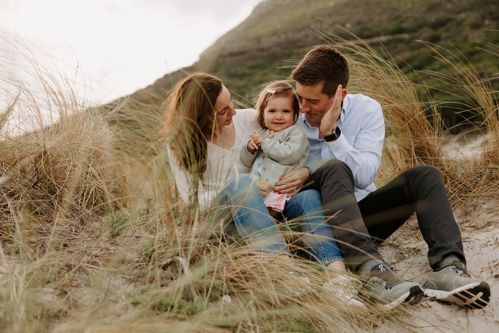 A happy family with a toddler sits together in coastal sand dunes surrounded by tall grass.