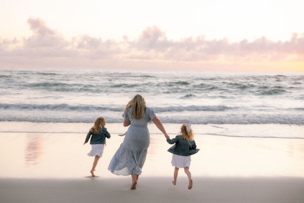 A mother holds hands with two young daughters walking toward ocean waves on a sandy beach at sunset.
