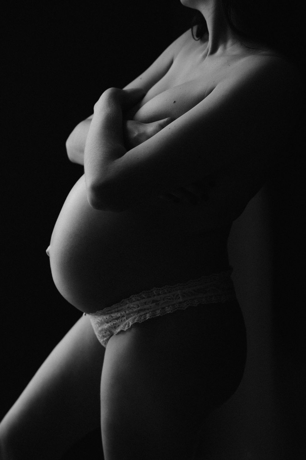 Black and white artistic maternity photo of pregnant woman cradling belly in soft dramatic lighting.