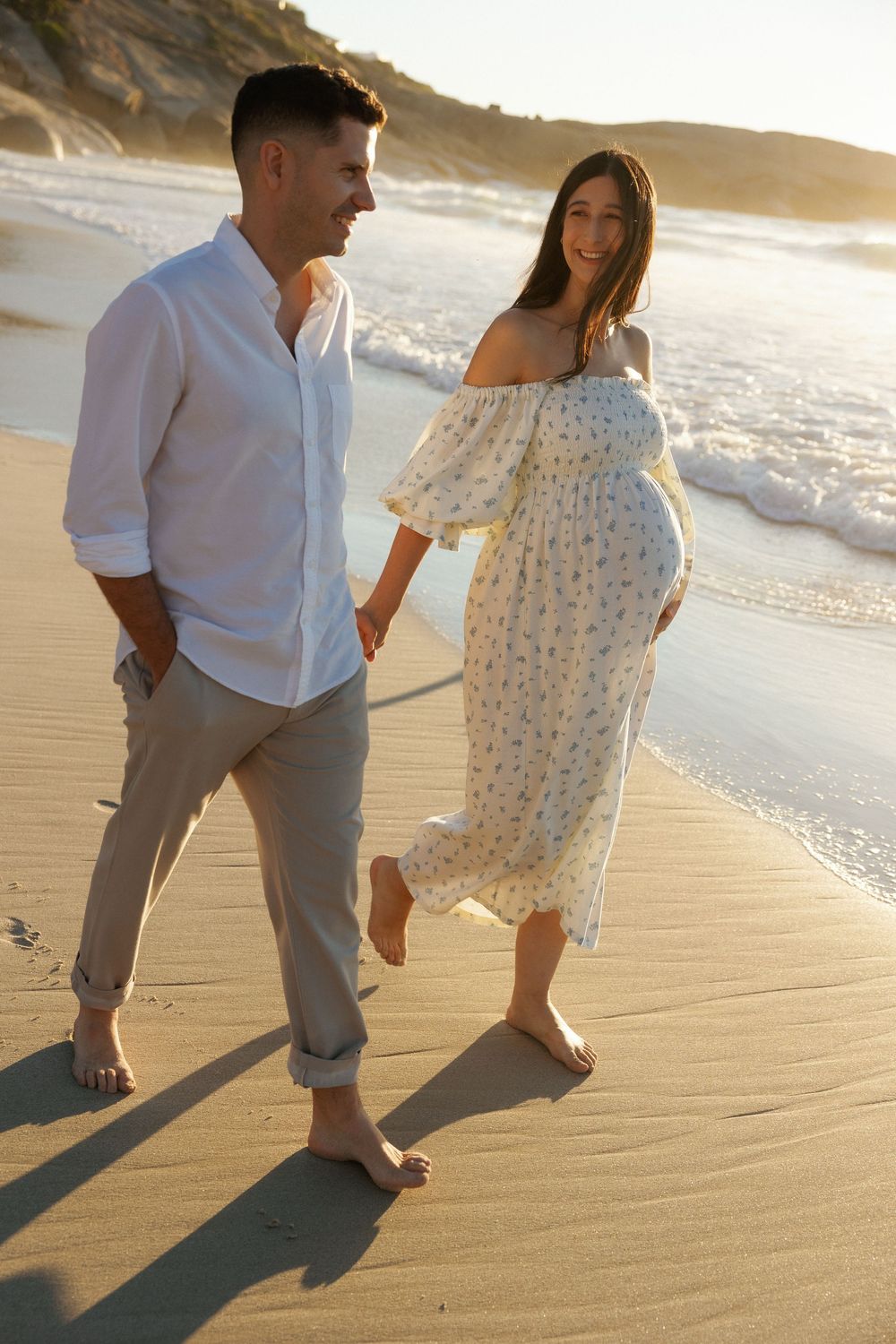 Pregnant couple walking barefoot on a sunny beach at golden hour, holding hands in casual summer outfits.