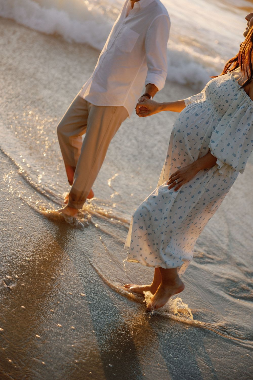 Pregnant couple walking barefoot on a sandy beach at sunset, holding hands as ocean waves wash over their feet.