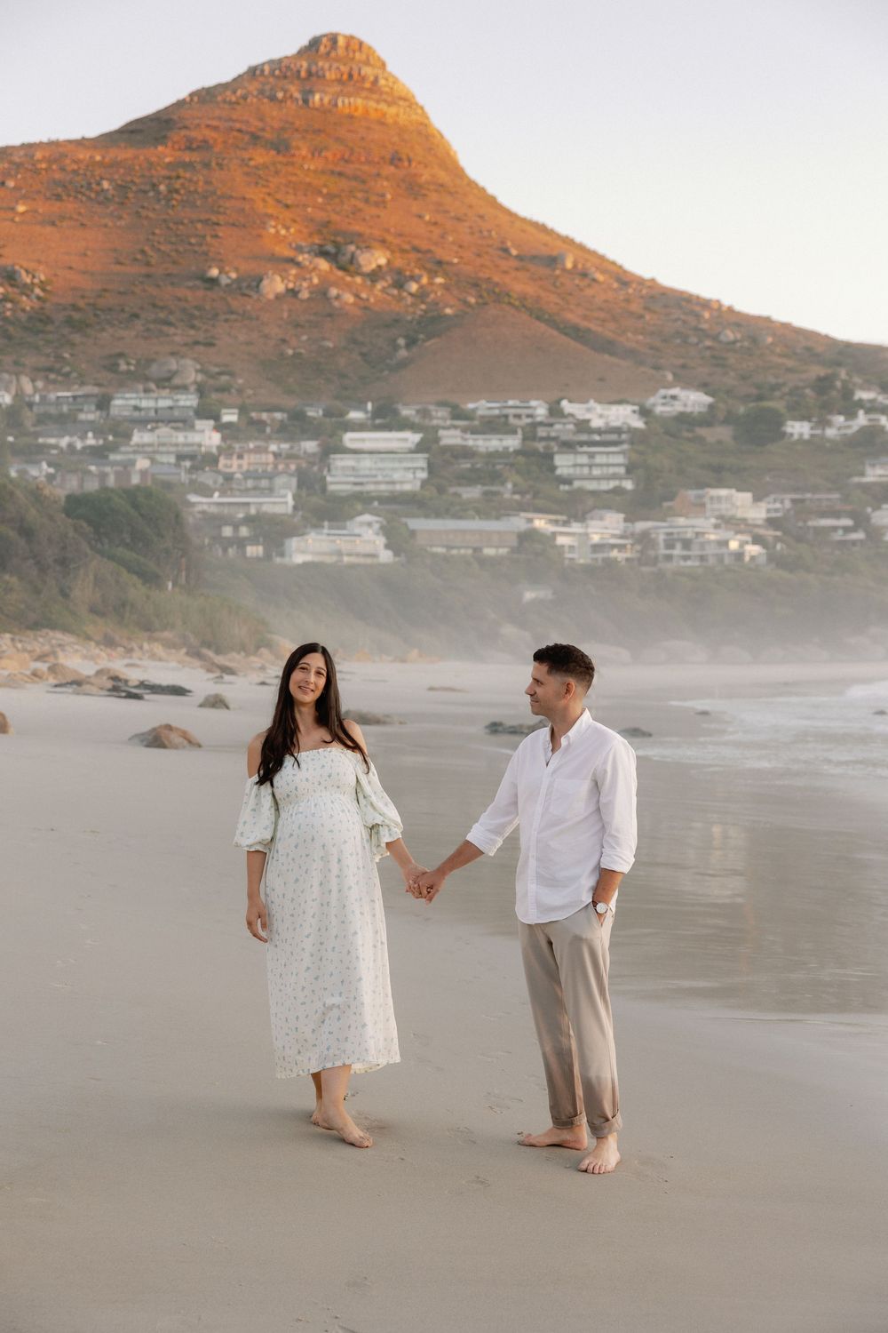 Couple holding hands on a sandy beach at sunset with Lion's Head mountain and coastal town in the background.